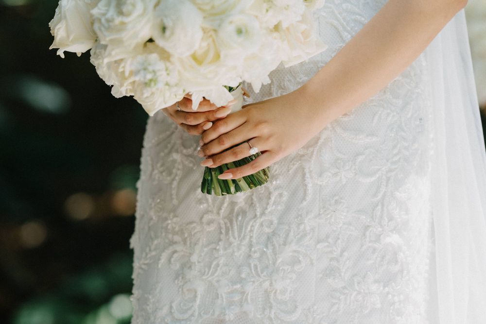 A close-up of hands holding a white rose bouquet against a lace wedding dress in soft natural lighting.