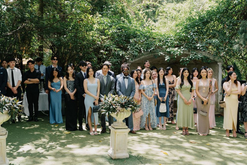A wedding party stands in line for photos beneath lush green trees on a sunny afternoon.
