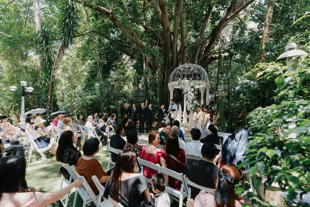 Guests gather at an outdoor garden wedding ceremony under a white gazebo surrounded by lush greenery.