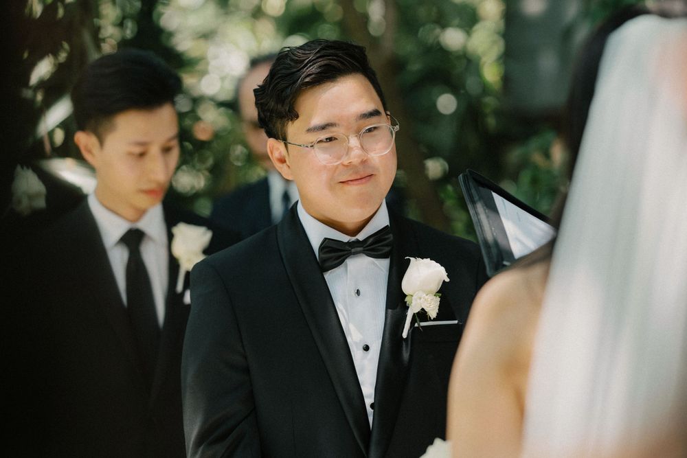 A groom in a black tuxedo with bowtie and white boutonniere smiles during an outdoor wedding ceremony.