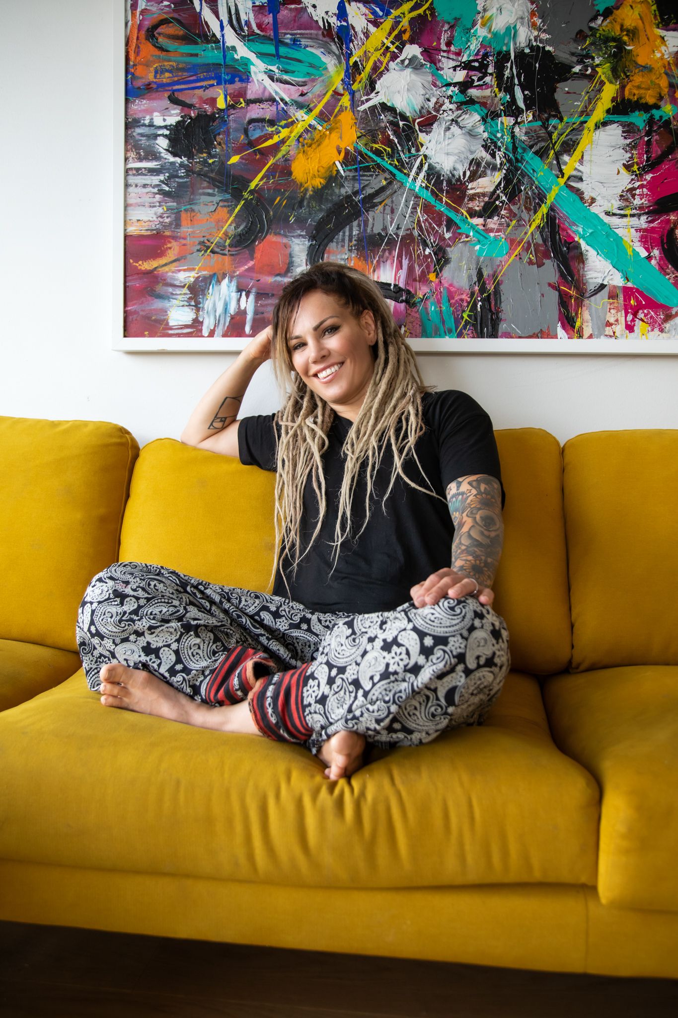 Cozy yellow sofa with black-clad figure in patterned pants relaxing beneath vibrant abstract wall art painting.