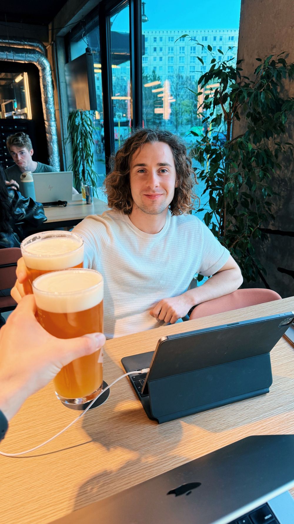 Person sits at cafe table with laptop and two tall glasses of beer while working near window with plants.