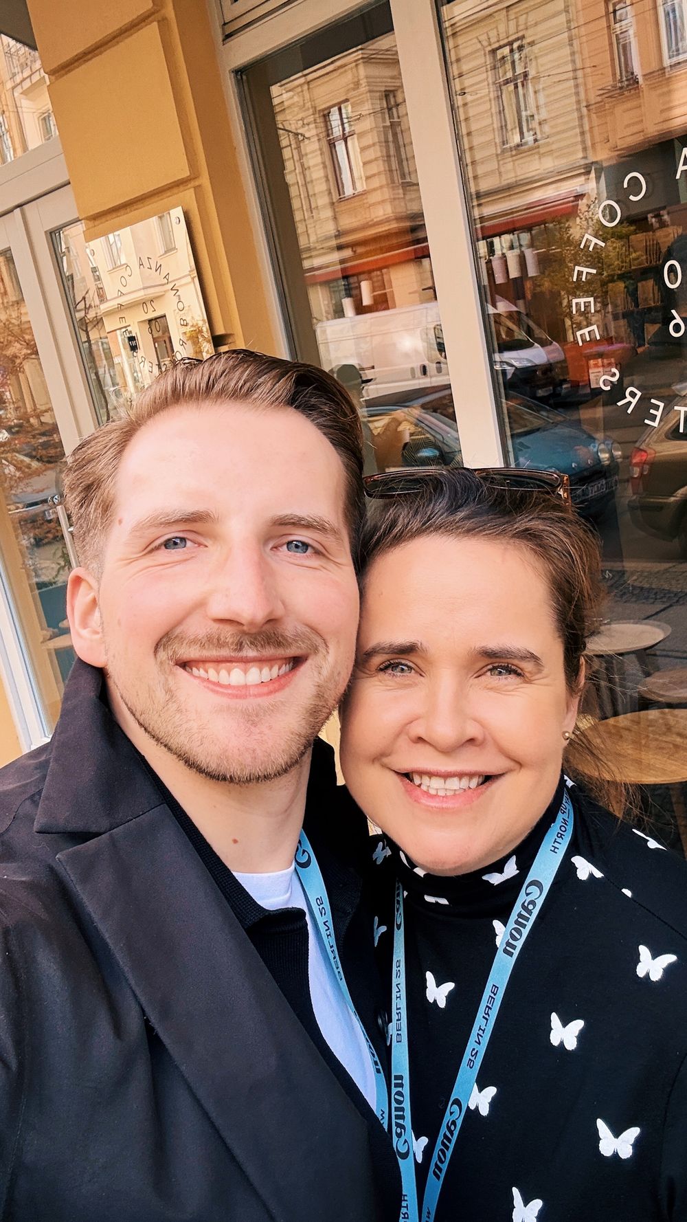 Two colleagues wearing lanyards take a selfie together outside a shop window on a sunny day.