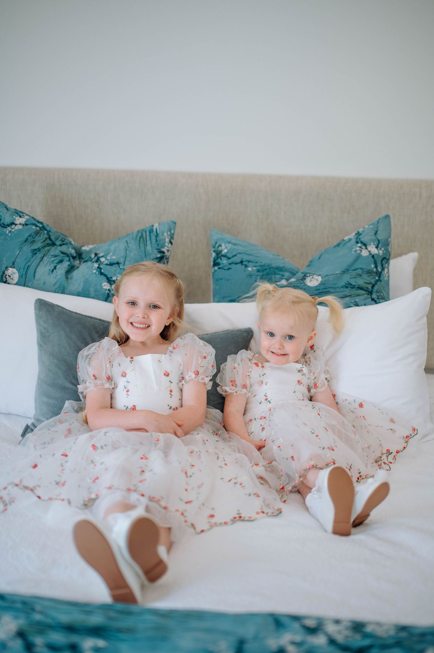Two young children in matching floral dresses sit together on a bed with teal pillows.