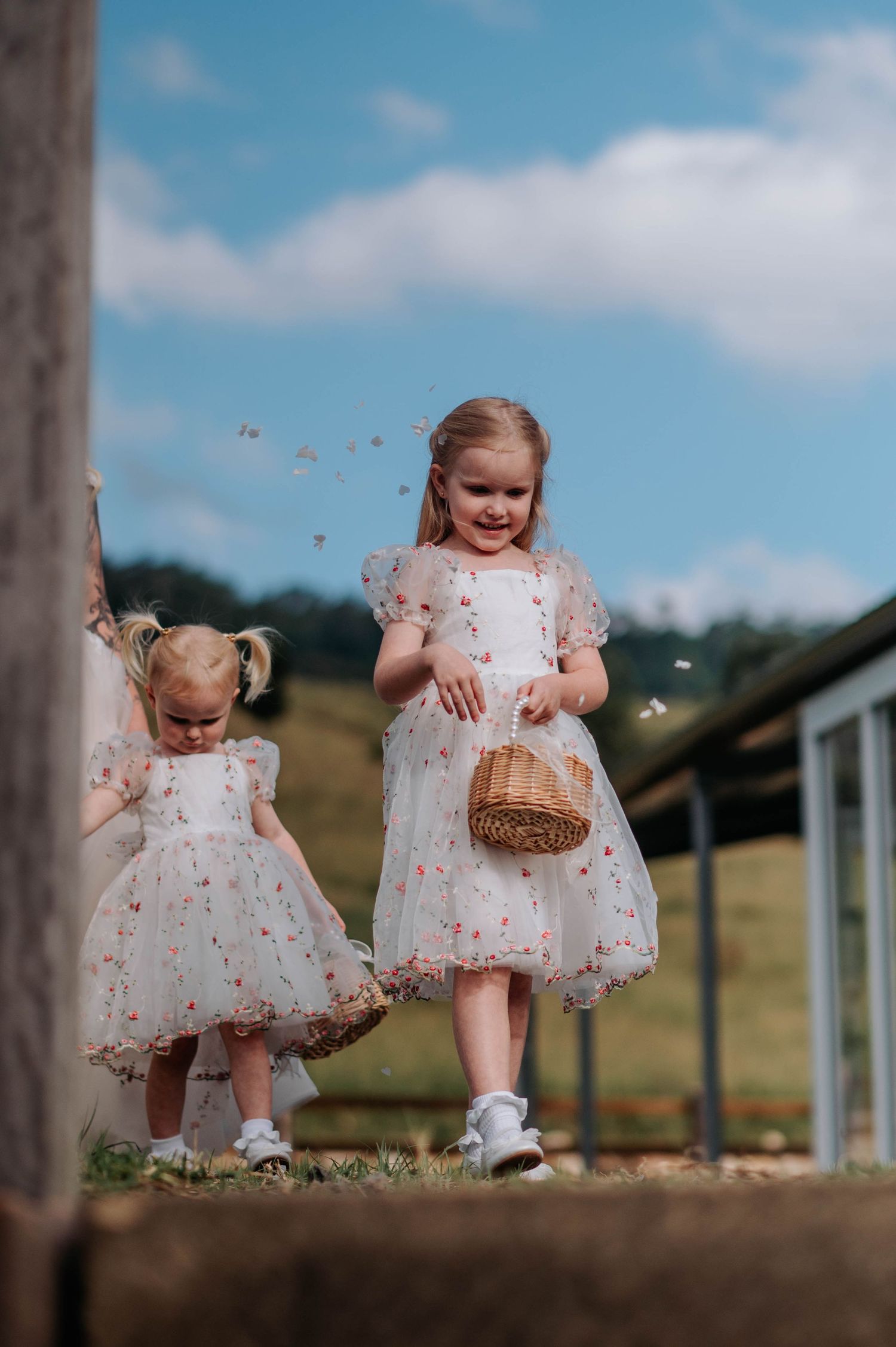 Children in white floral dresses play outdoors with a small basket on a sunny day.