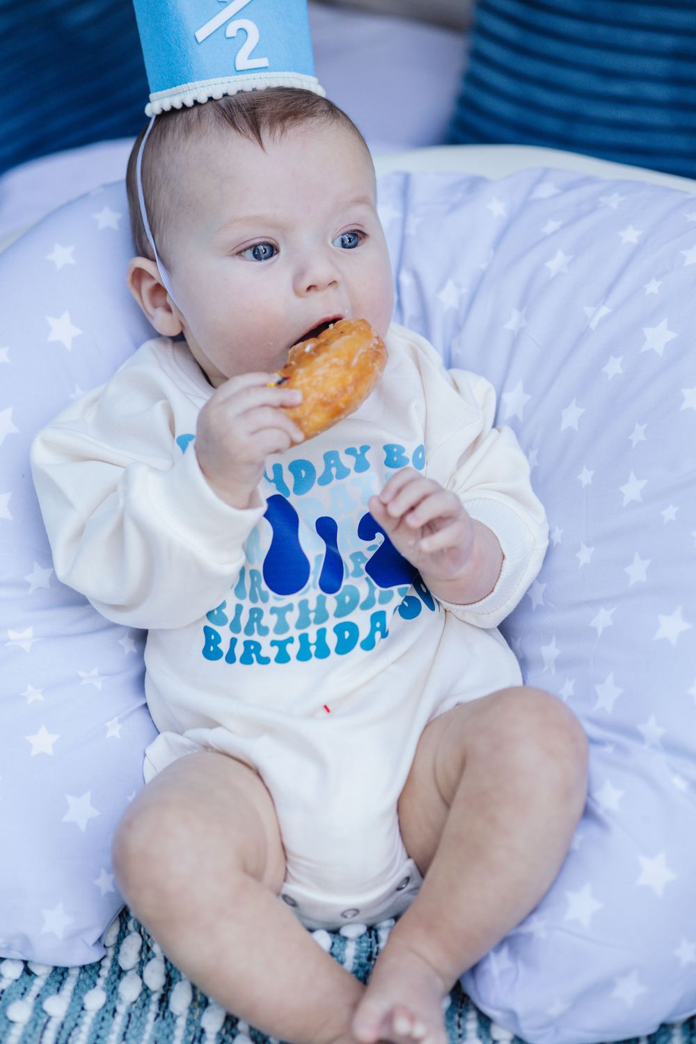 Baby in white birthday onesie with blue lettering lies on star-patterned bedding while eating a cookie.