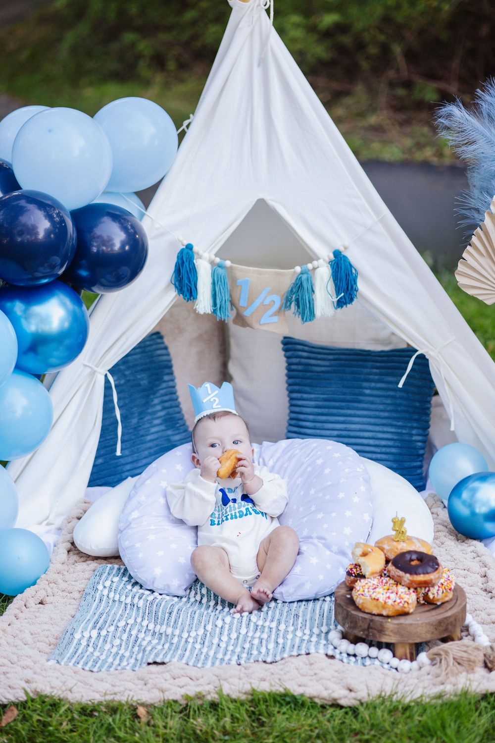 Baby photoshoot setup with white teepee tent, blue balloons and blankets on grass creating a dreamy outdoor scene.