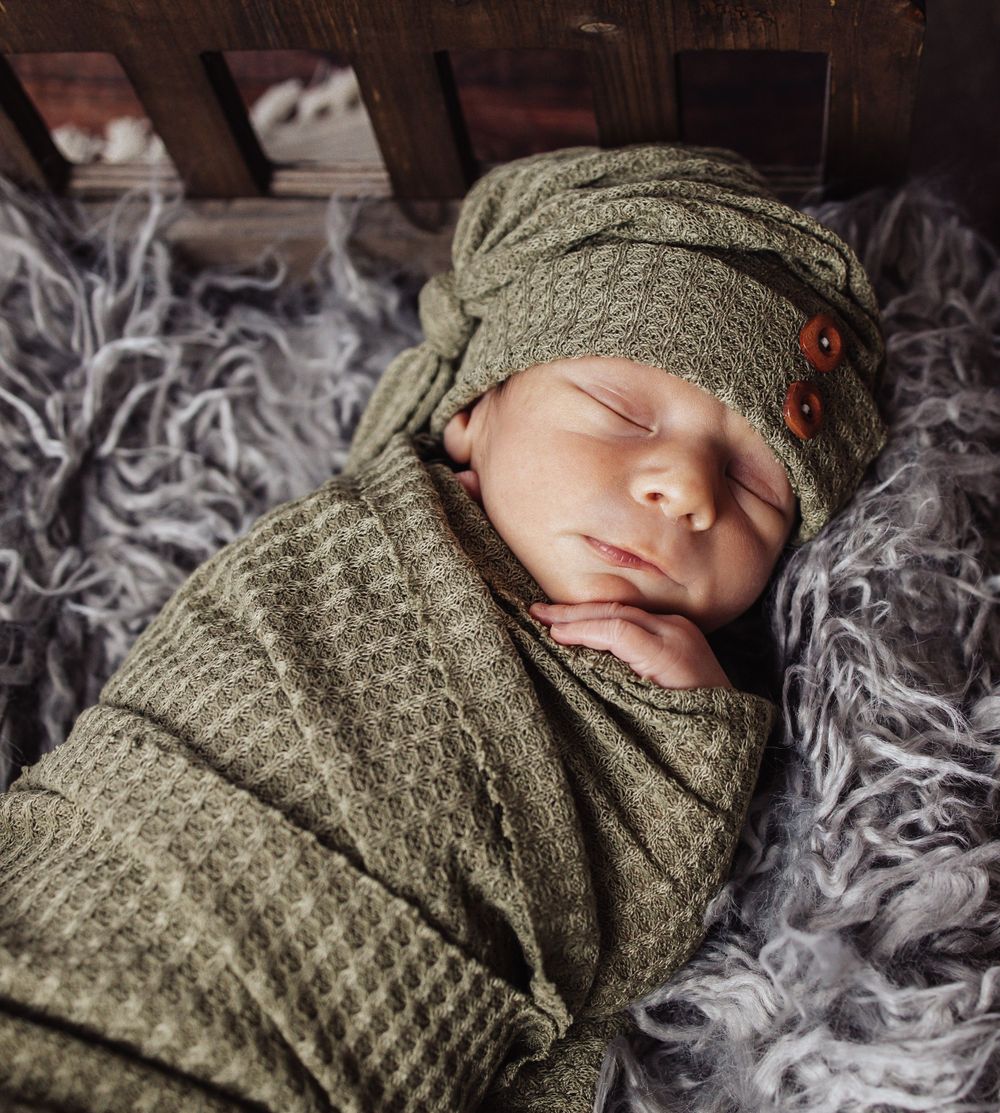 Sleeping newborn wrapped in olive green knitted swaddle and matching hat rests on soft gray fur blanket.
