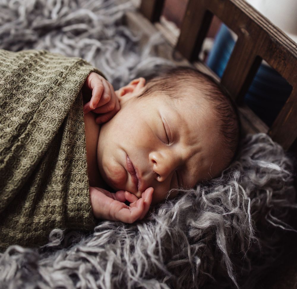 Peaceful sleeping newborn baby wrapped in olive green blanket rests on soft gray fur.
