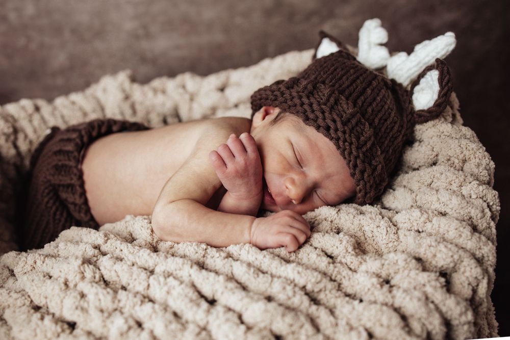 Sleeping newborn wearing brown knit hat with antlers on textured beige blanket.