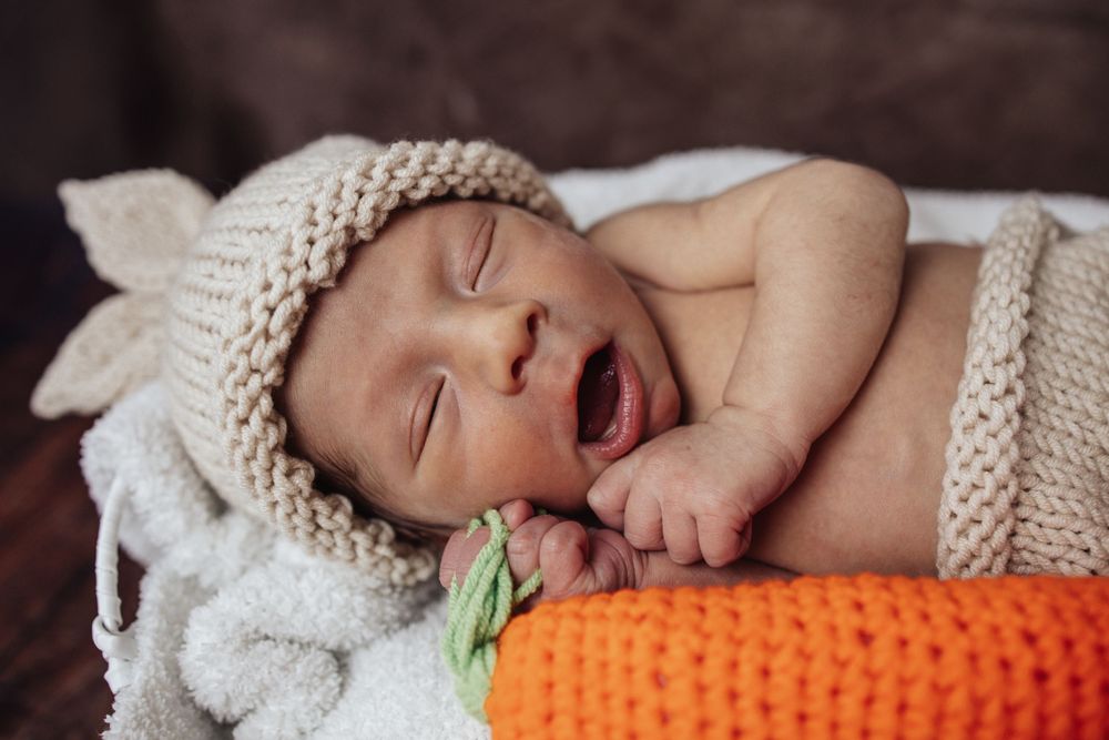 Sleeping baby in knitted beige hat lies next to orange crocheted carrot toy on white blanket.