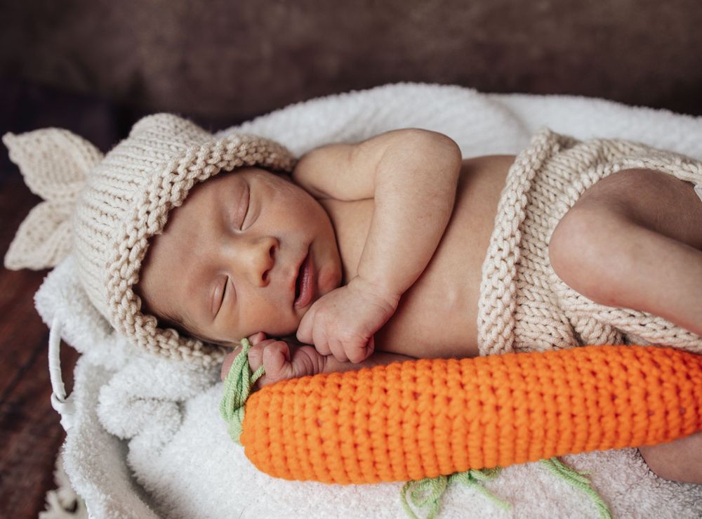 Sweet newborn sleeping next to a knitted carrot toy while wrapped in cozy white blanket.