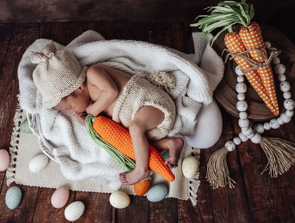 A cozy newborn photography setup with wooden beads, knit blankets, and decorative carrot props arranged on dark wood flooring.