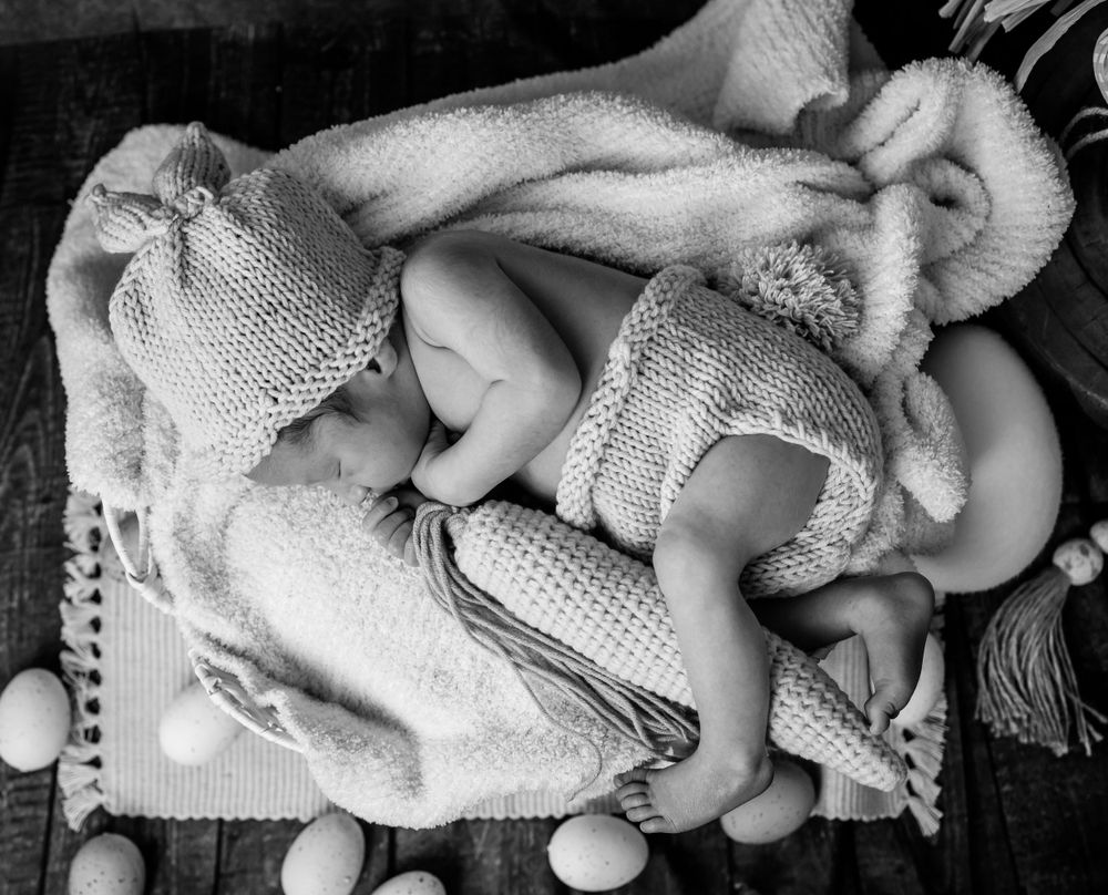 Two babies swaddled in white knitted blankets sleeping together on a textured surface.