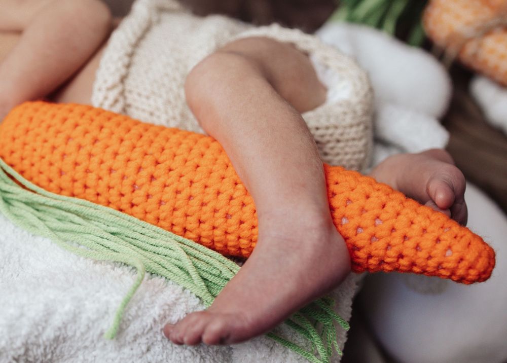 Baby wearing crocheted orange carrot costume bootie accessory lies on white blanket.