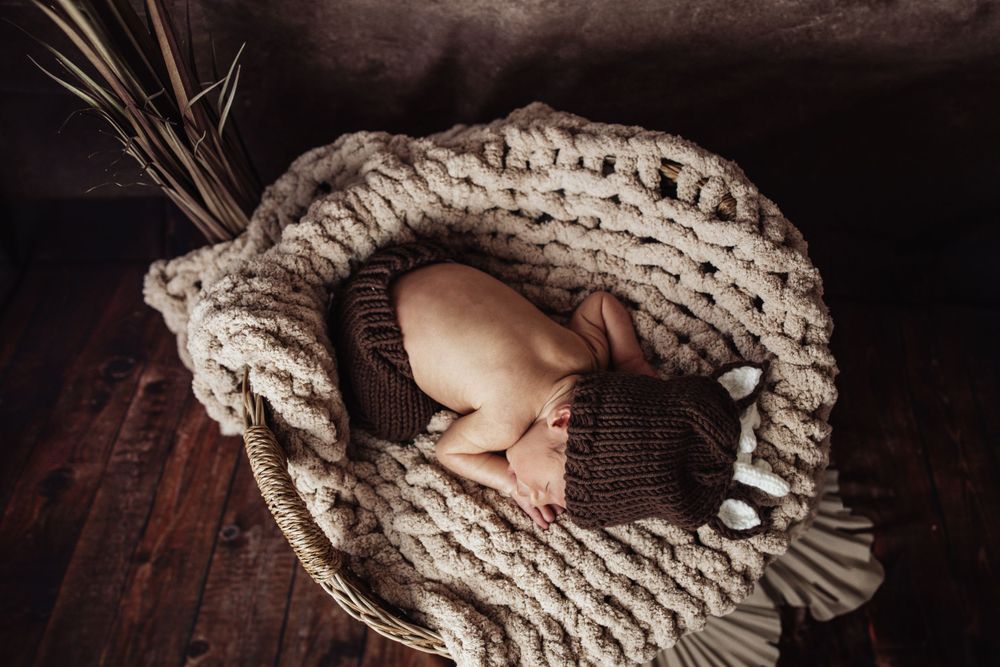Newborn sleeping wrapped in thick knitted blanket on dark wooden floor.