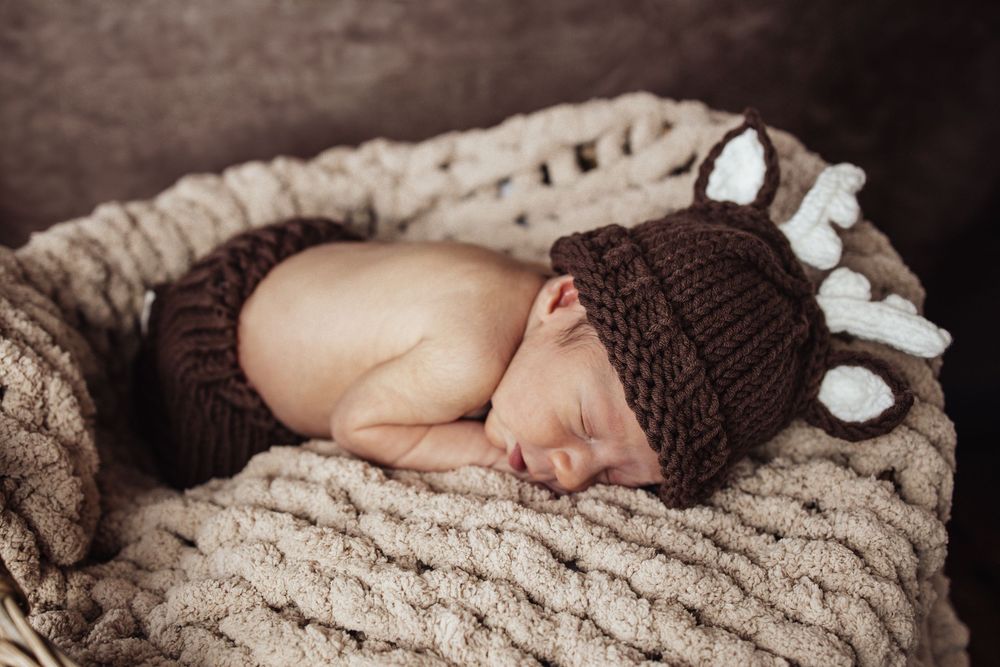 Peaceful sleeping newborn wearing a brown knitted hat rests on cozy beige textured blanket.