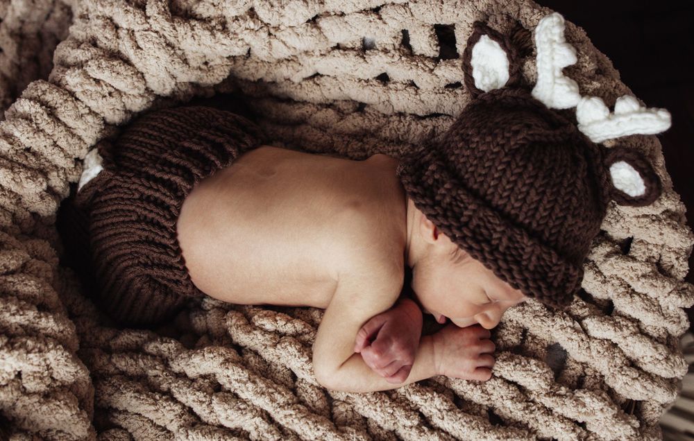 A sleeping infant wearing a brown knitted outfit curls up on a textured beige blanket.