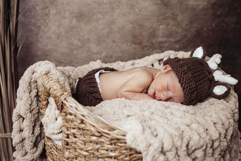 Newborn baby sleeping peacefully in brown knit hat on cream-colored textured blanket in rustic basket.