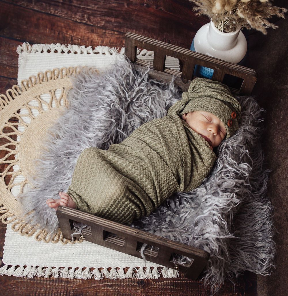 A newborn sleeps peacefully in a rustic wooden basket lined with fluffy gray fur and a cream doily.