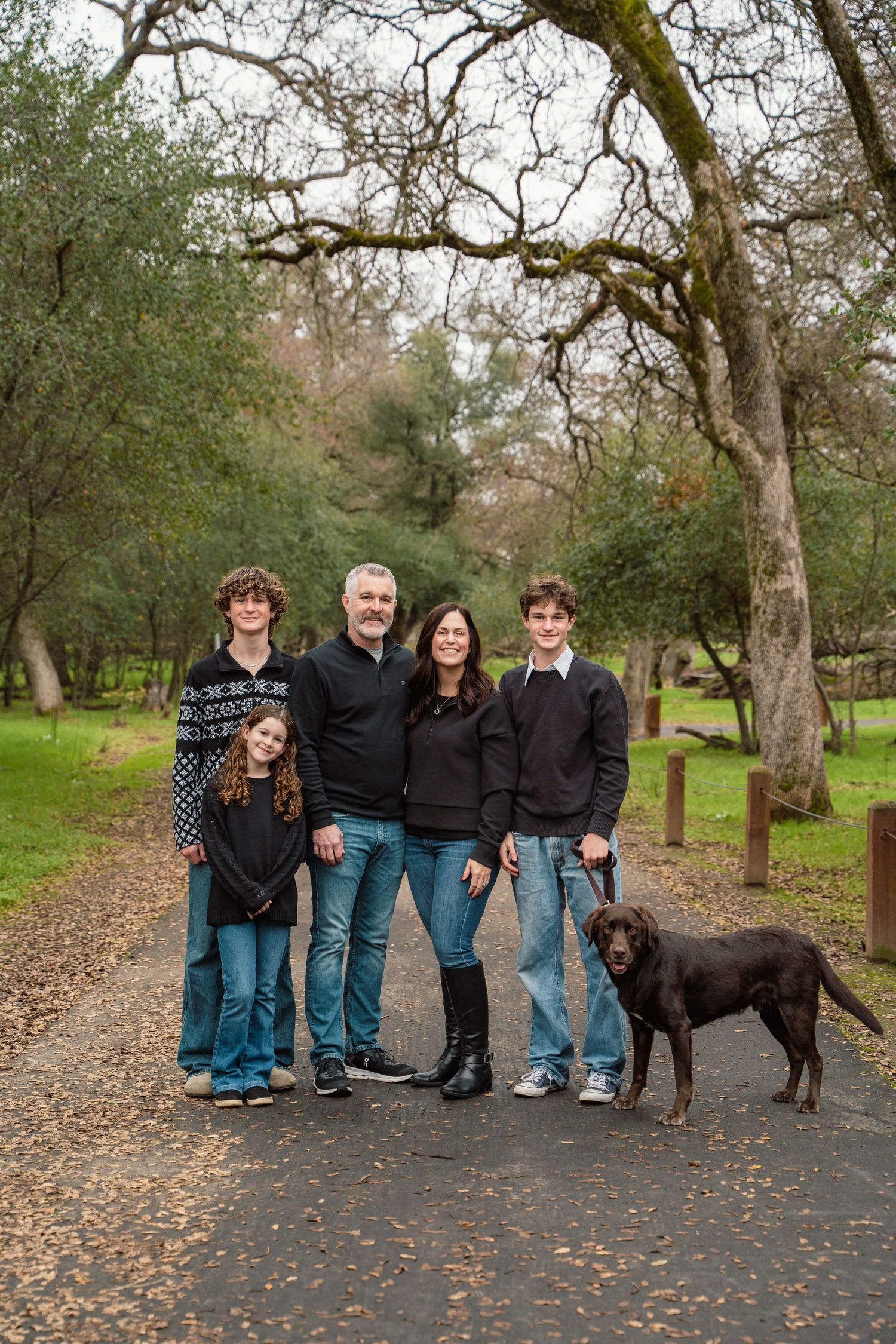 A family of five poses with their black dog on a tree-lined path during an outdoor photoshoot.