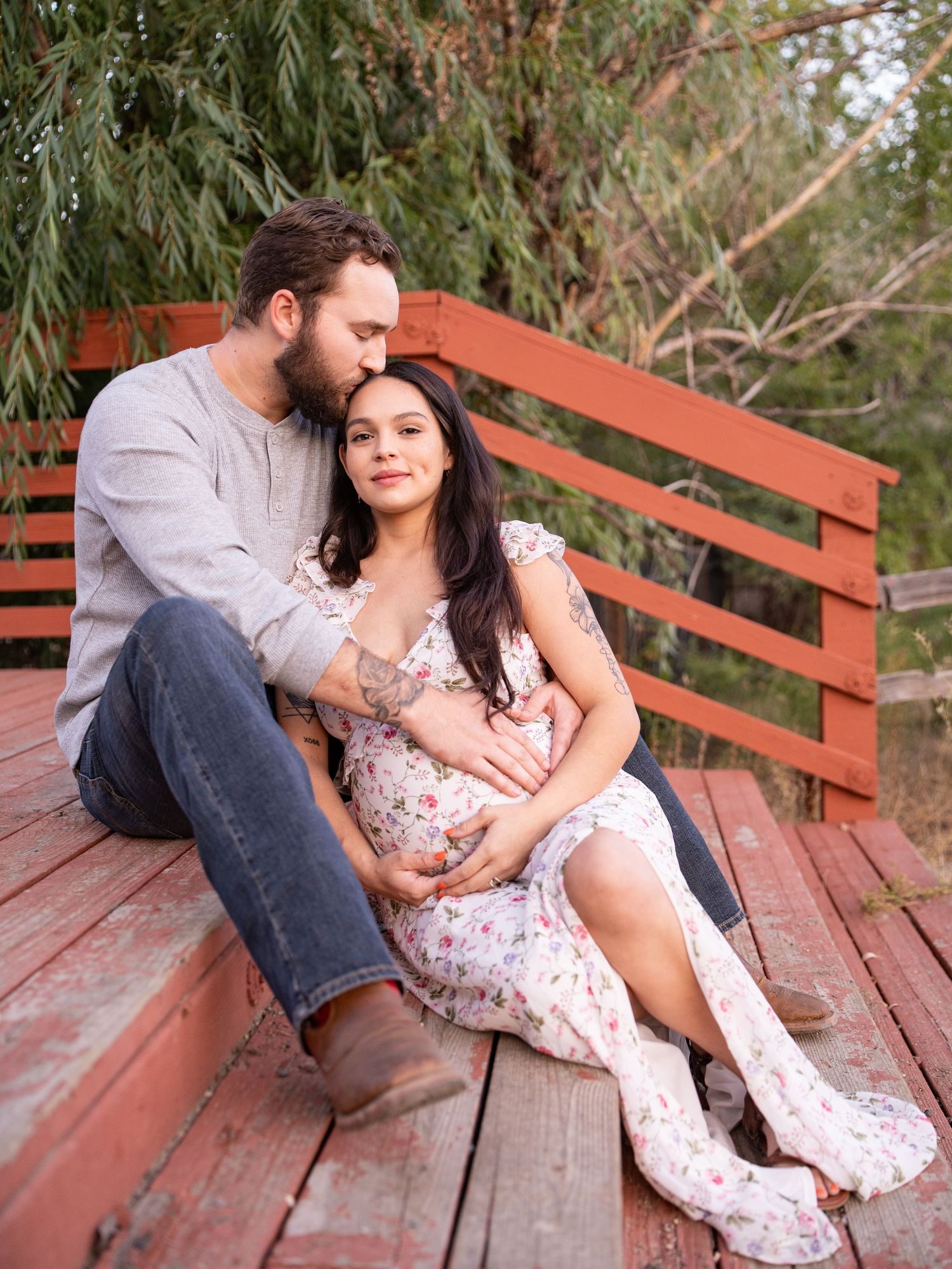 A couple relaxes together on a rustic red wooden bench surrounded by willow trees in a romantic outdoor setting.