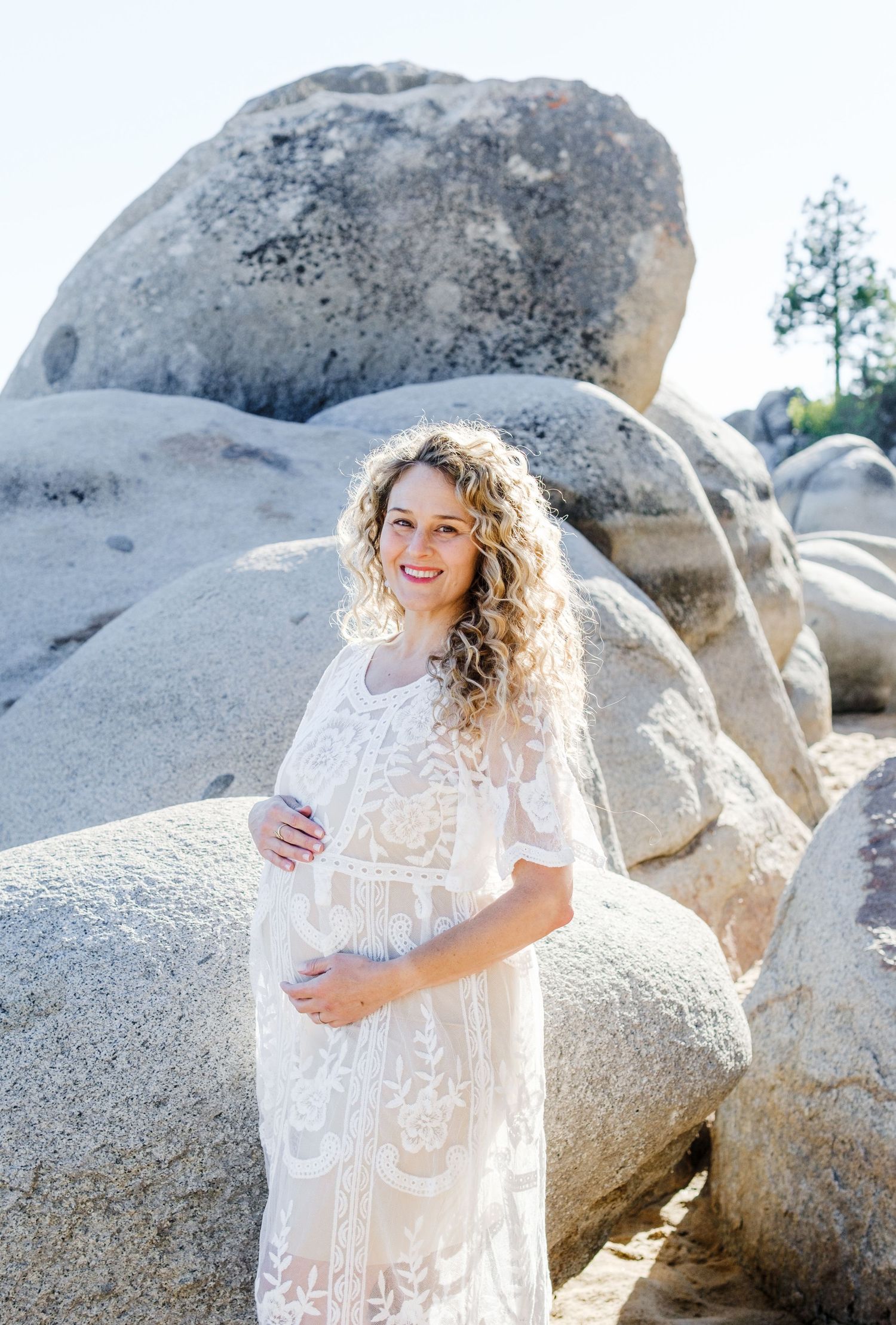 Expecting mother in white lace dress poses against large granite boulders in sunlit natural setting.