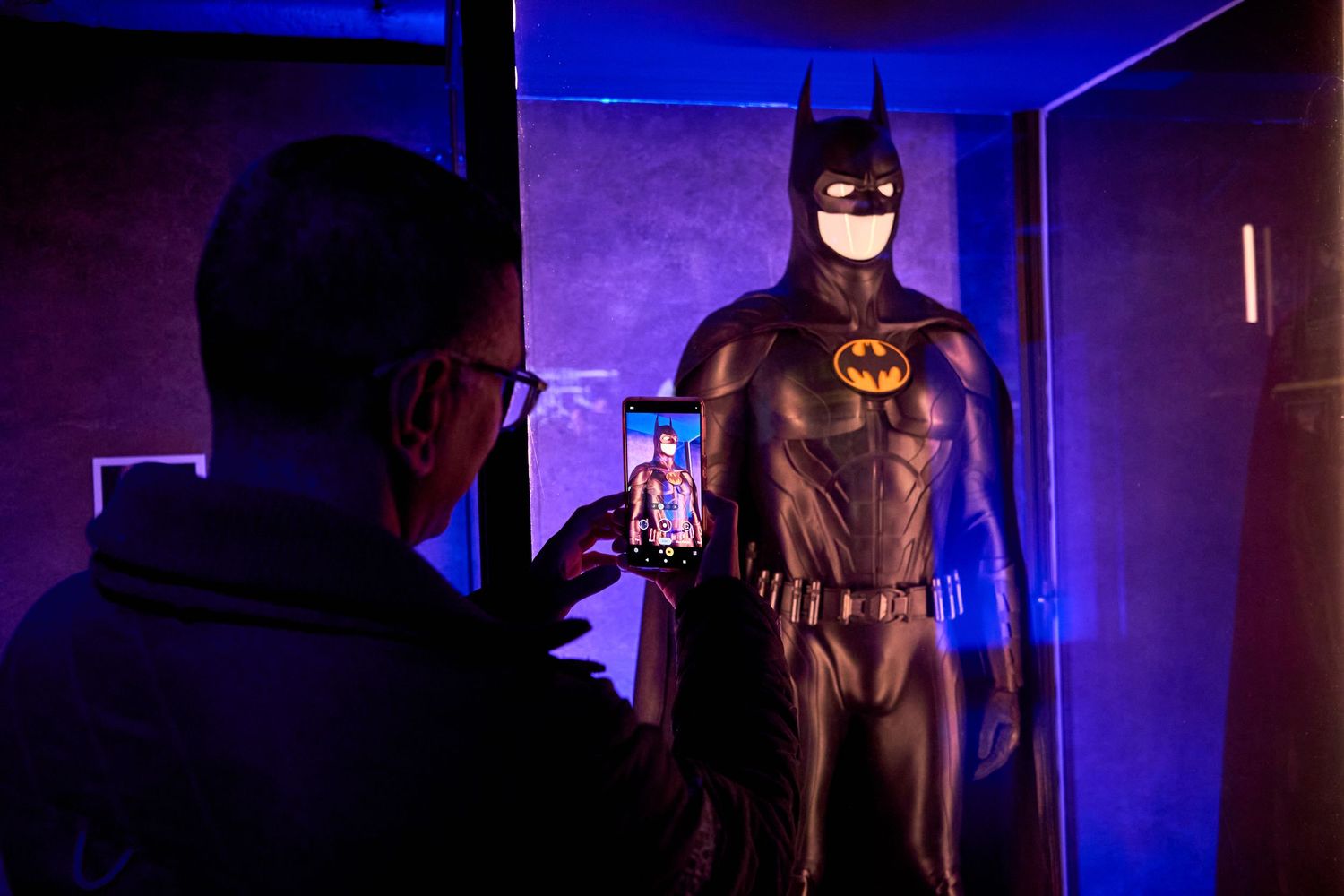 Batman costume display in dark room with purple lighting and someone taking a photo.