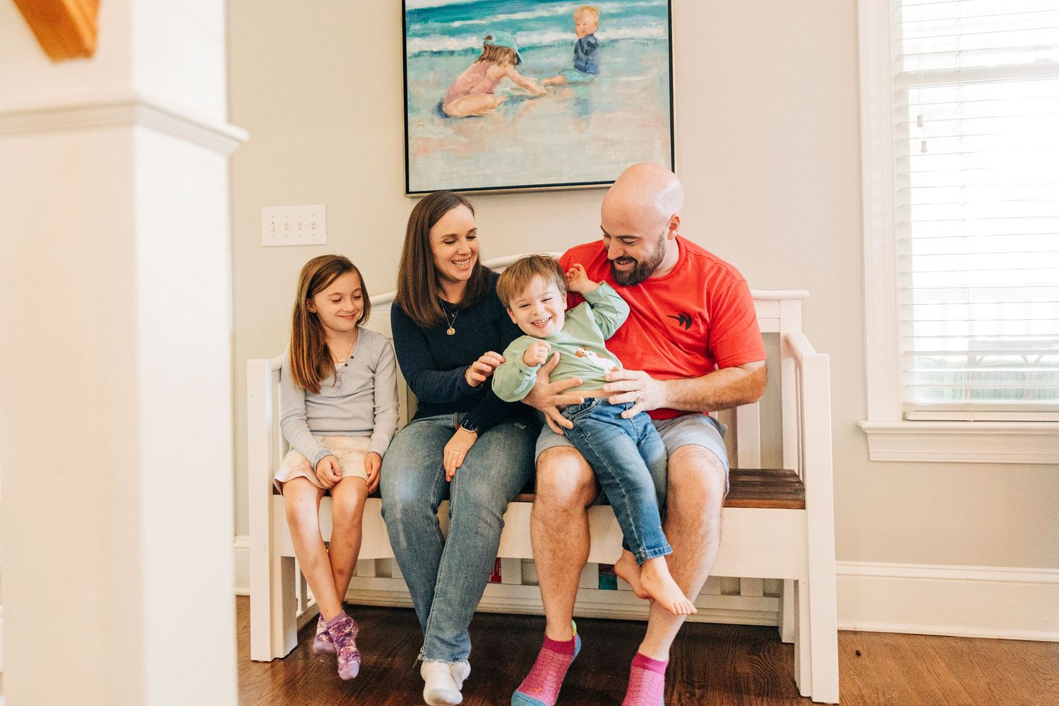 A family wearing colorful socks sits together on a white bench in a bright room with a beach painting on the wall.