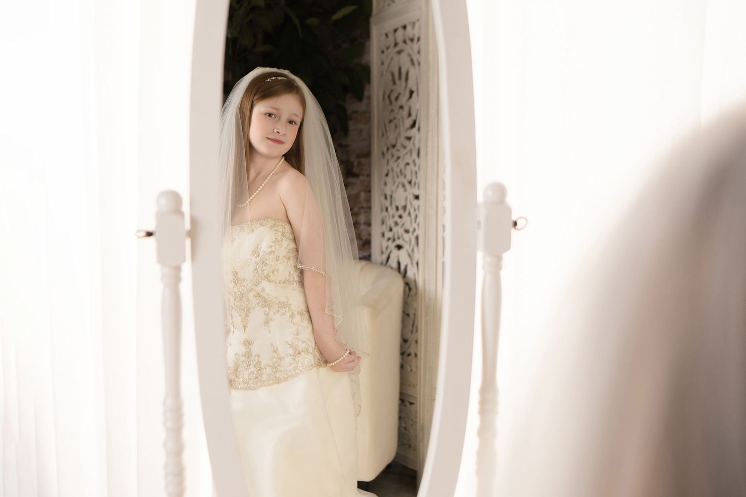A bride in a delicate lace wedding dress and veil stands elegantly in a bright white hallway.