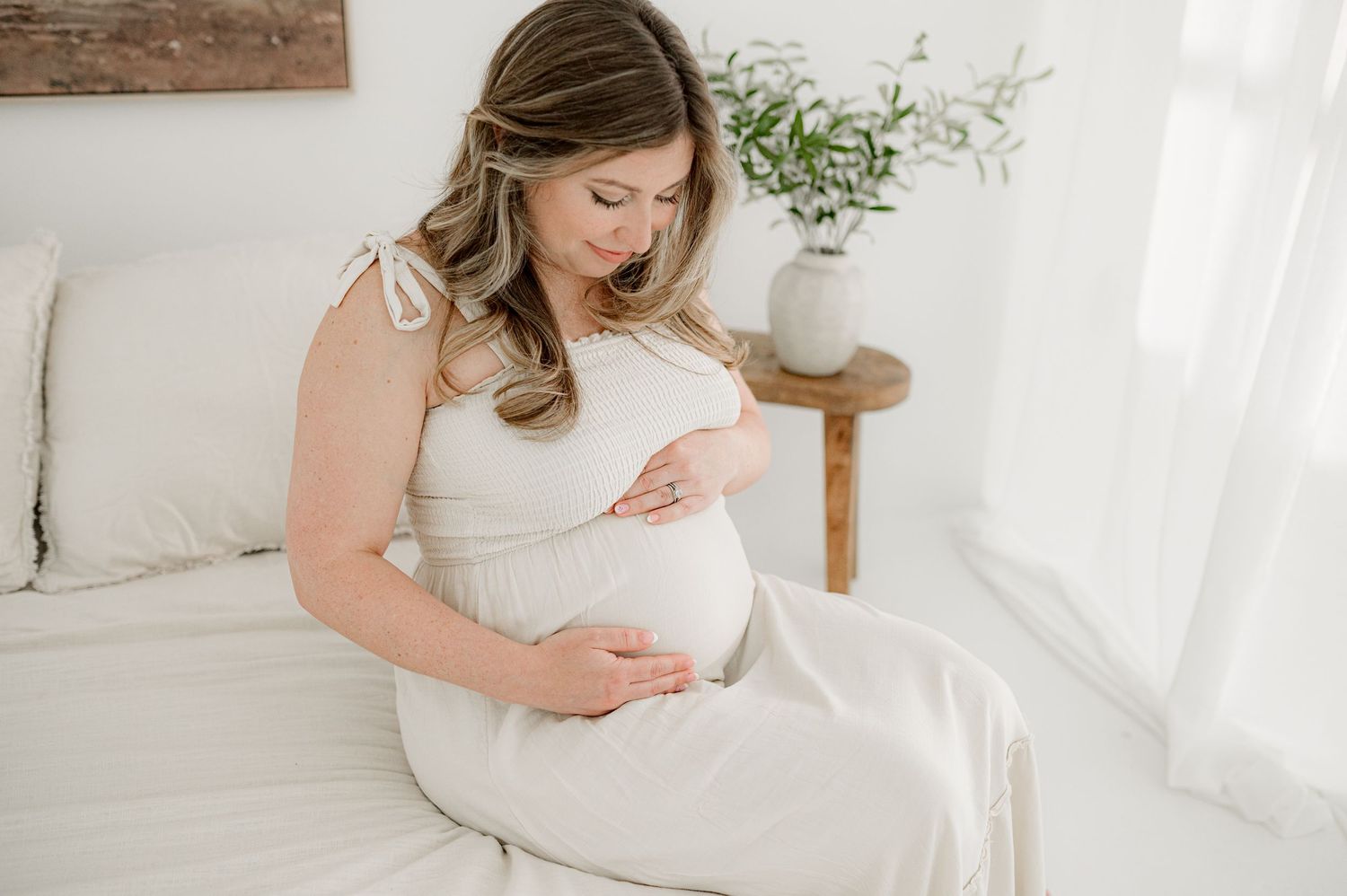 A pregnant woman in a white dress sits on a bed in a bright, airy room during her maternity photoshoot in Jacksonville.