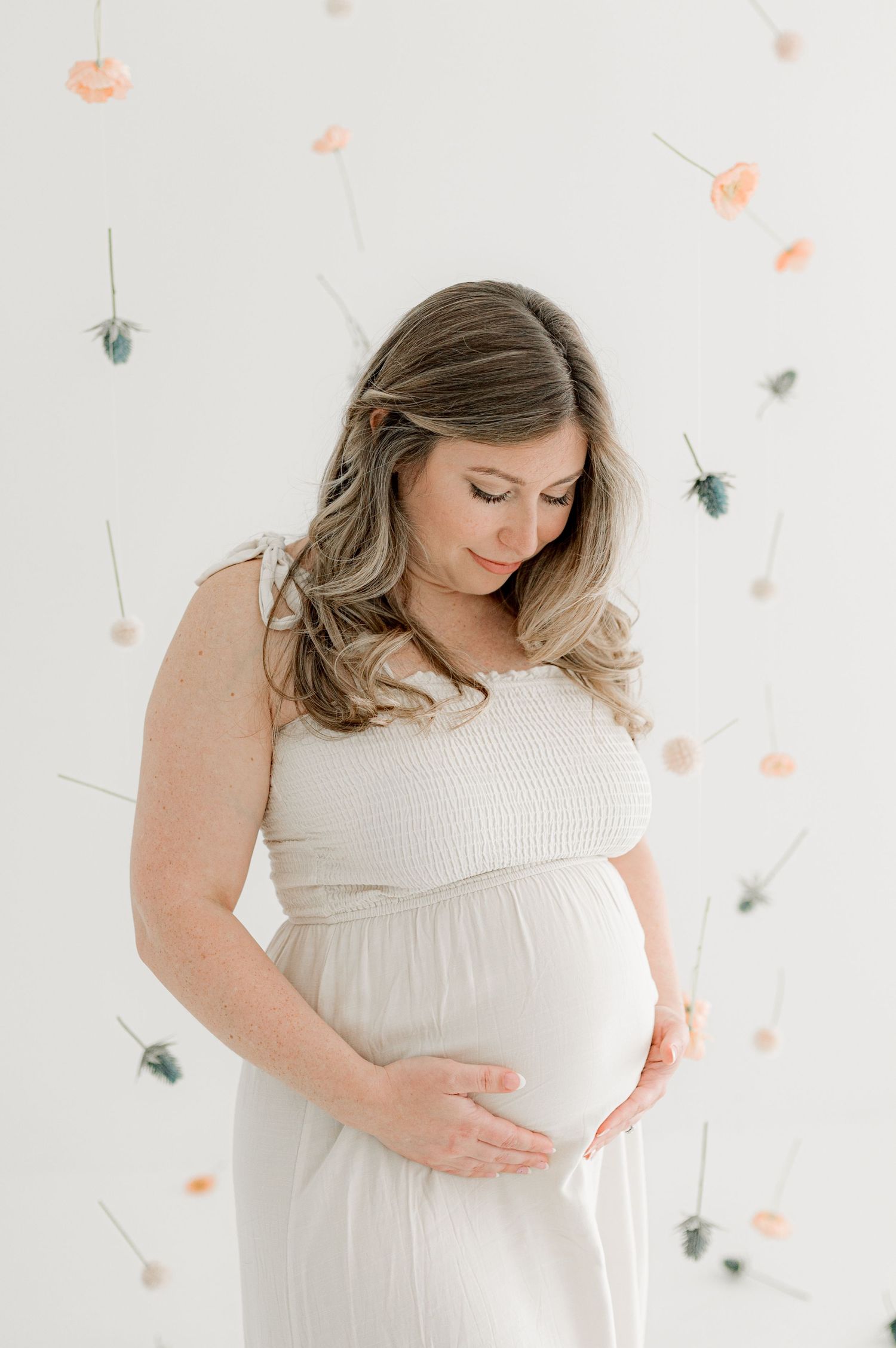 A pregnant woman in a white dress poses against a floral backdrop during her maternity photoshoot in Jacksonville, Fl.