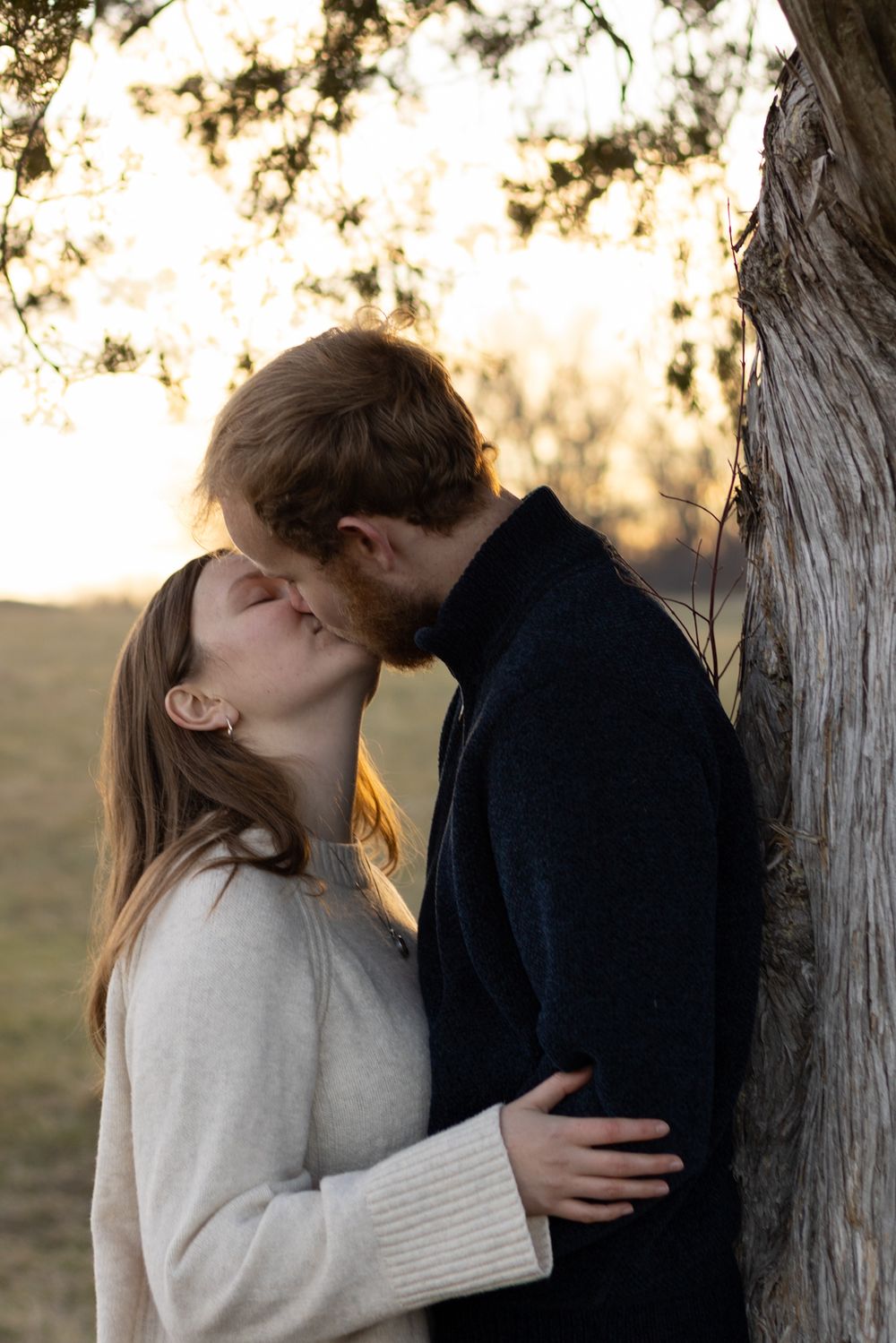 Couple shares intimate moment near tree trunk during golden hour.