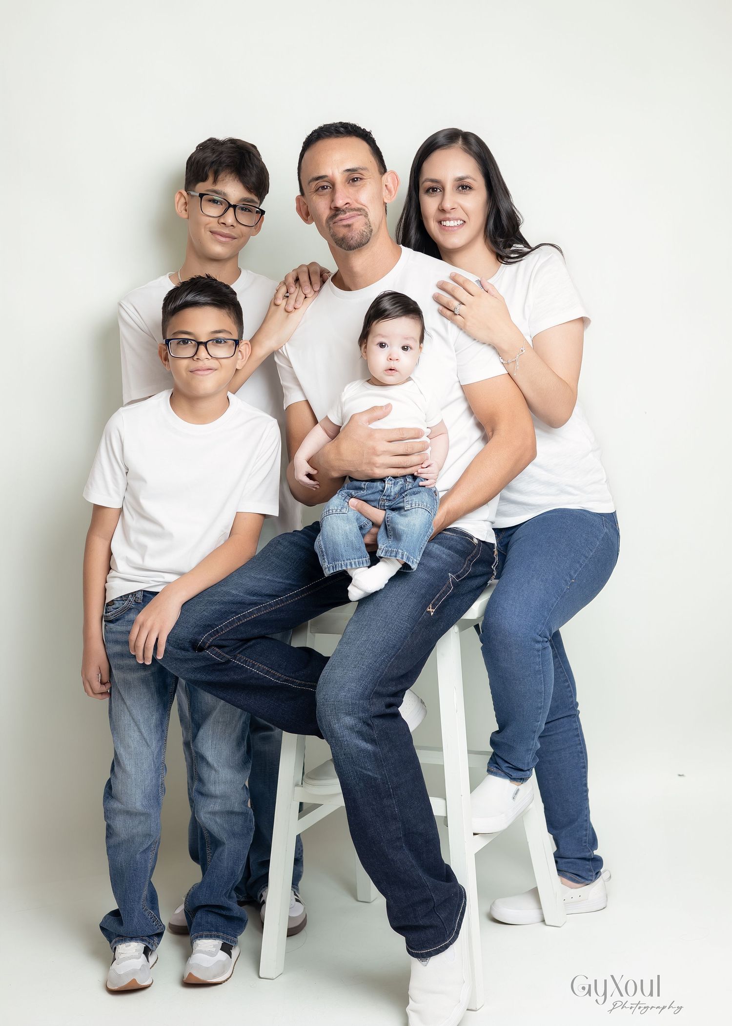 A family wearing white tops and jeans poses together against a light background in a studio setting.