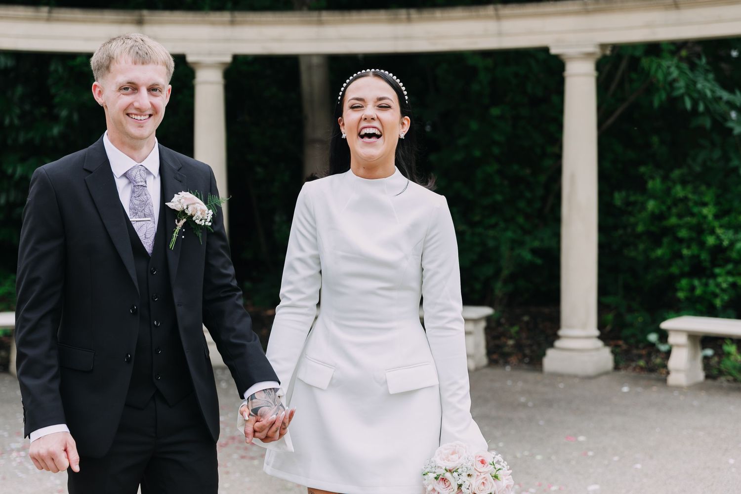 Wedding guests throw confetti at a newlywed couple exiting under a classical stone gazebo.