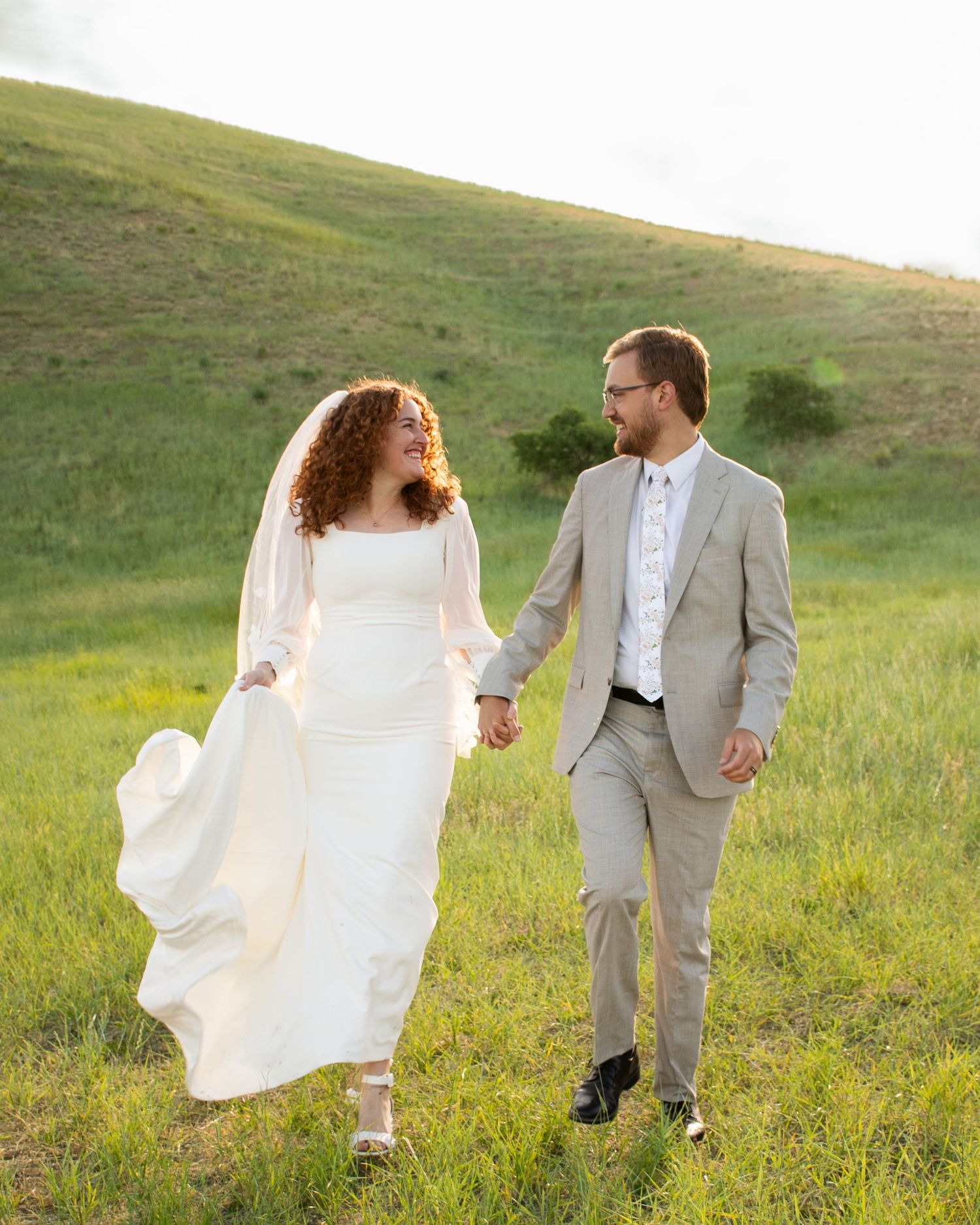 A couple walks together through a sunlit meadow of tall grass during a wedding photo session.