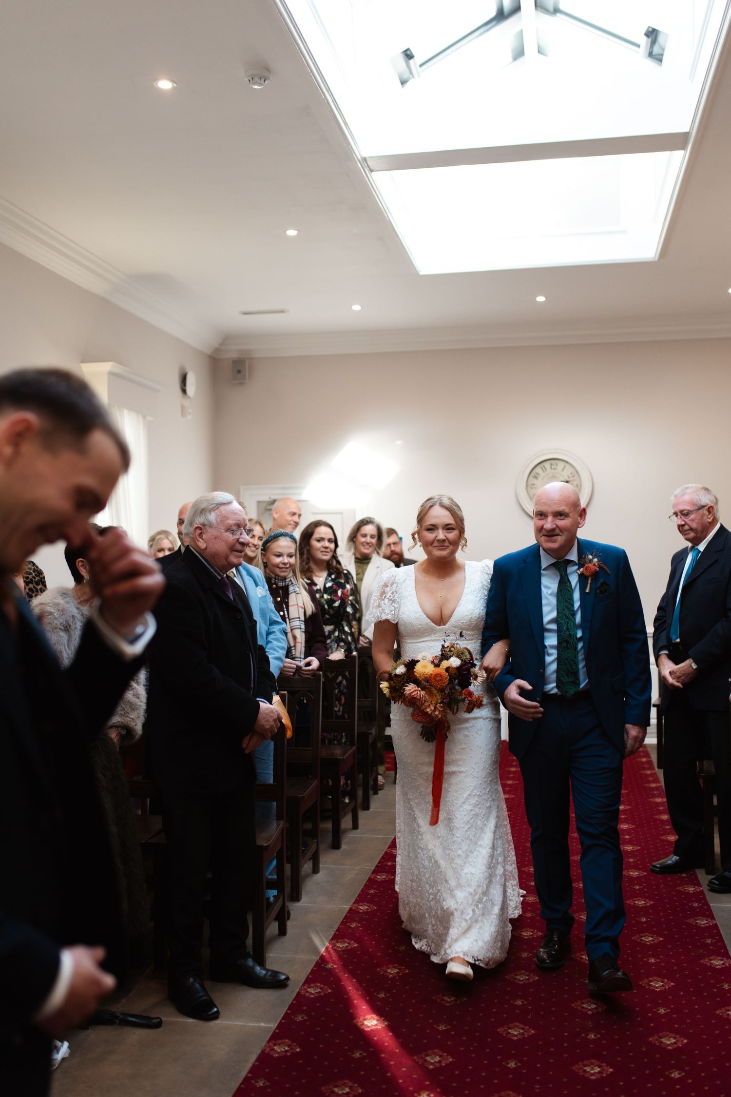 Wedding guests line a red-carpeted aisle as the bride walks down with her escort during an indoor ceremony.