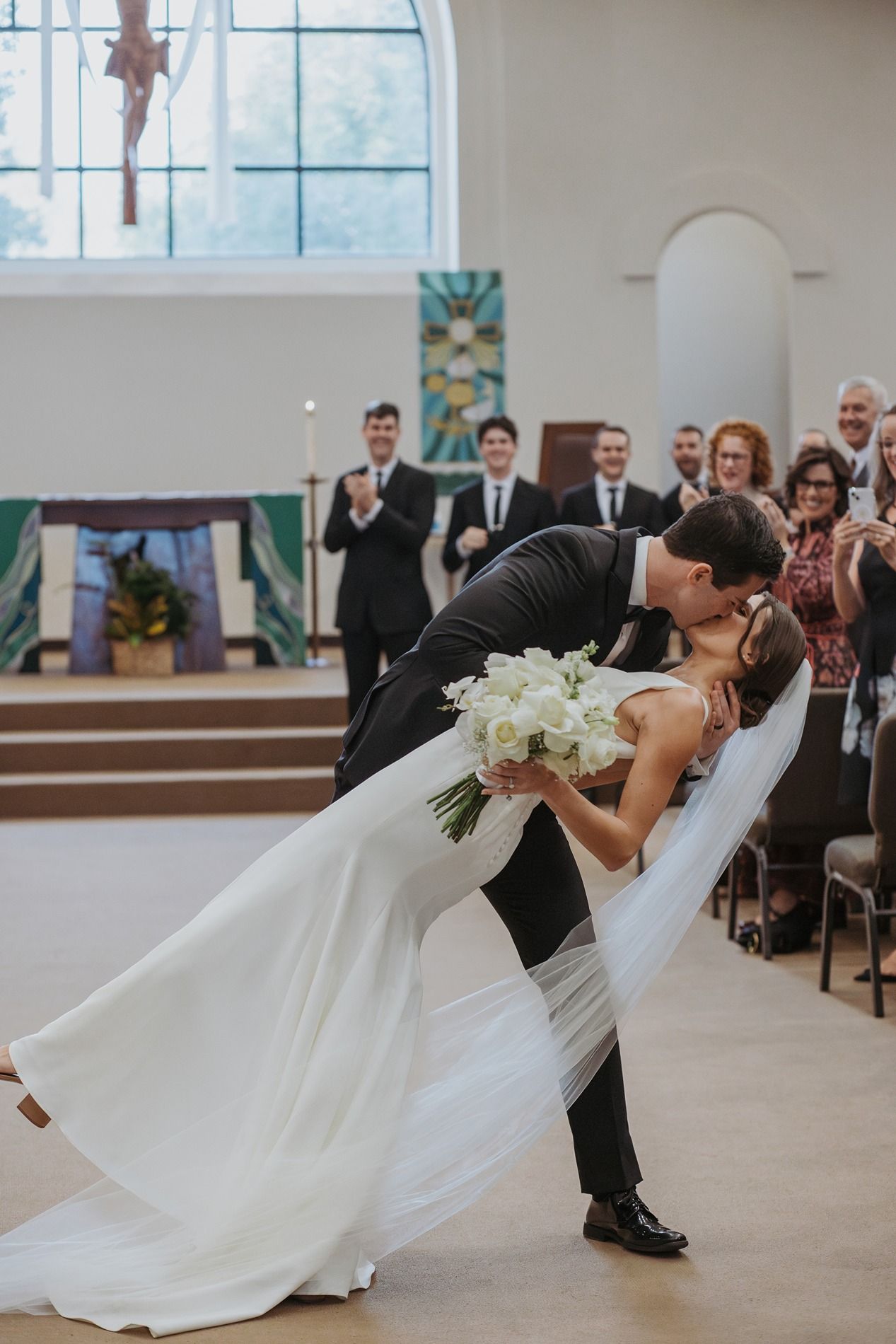 A couple performs a romantic dip during their wedding ceremony in a bright church venue.