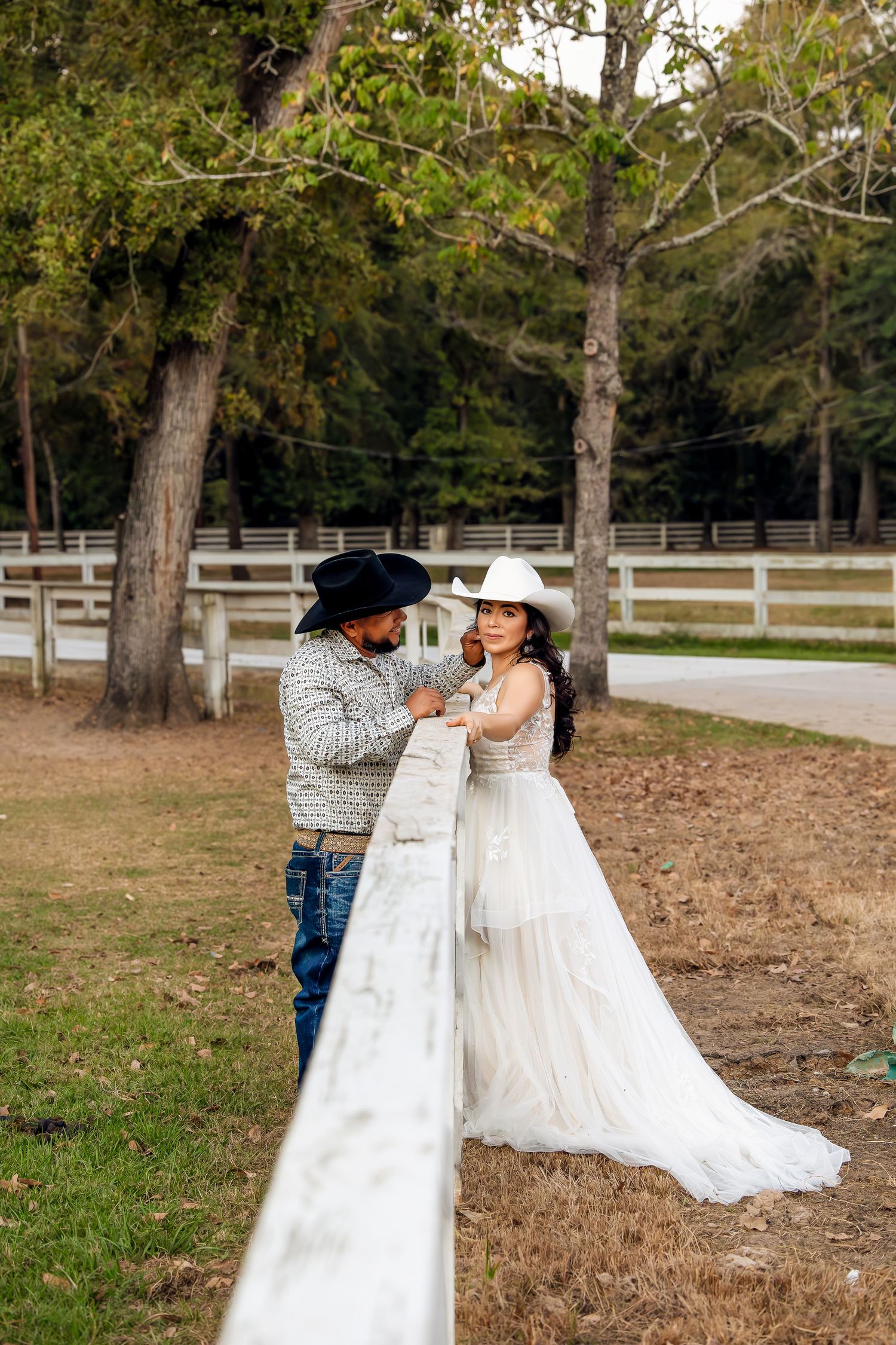 A couple in western attire dances together in a rustic outdoor setting with white fencing and autumn trees.