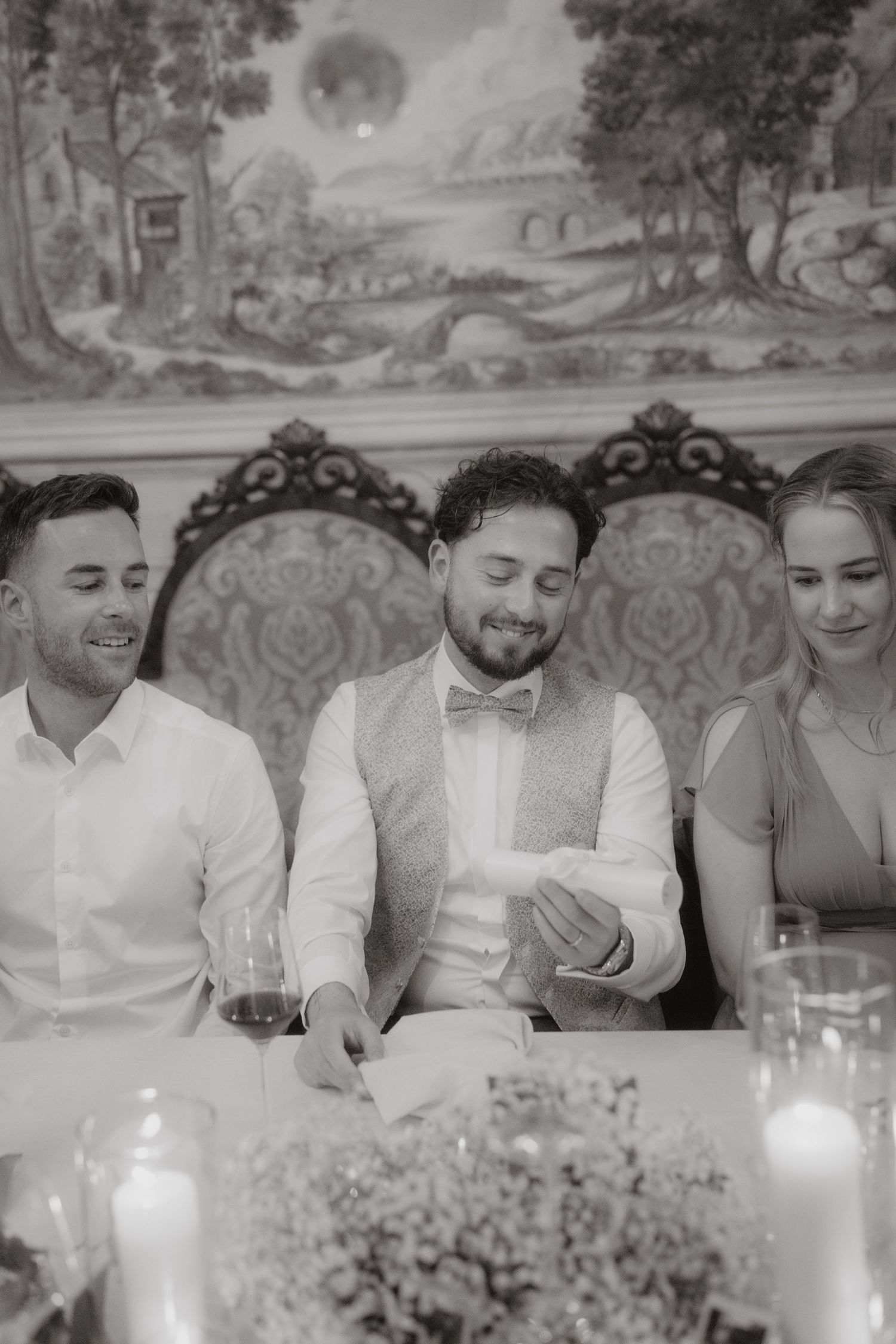 Guests seated at a formal dinner table with decorative wallpaper in the background.