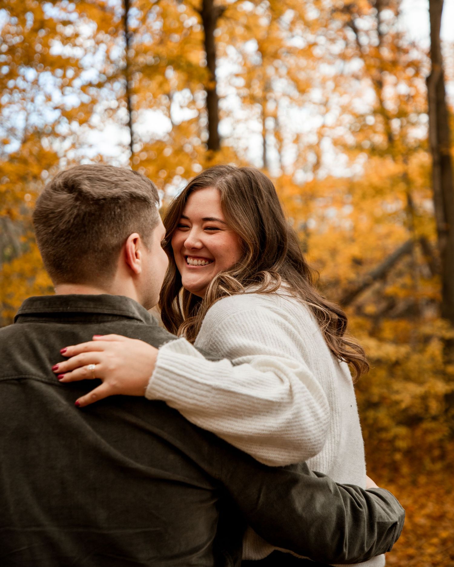 A couple embraces joyfully in a forest with vibrant orange autumn foliage in the background.
