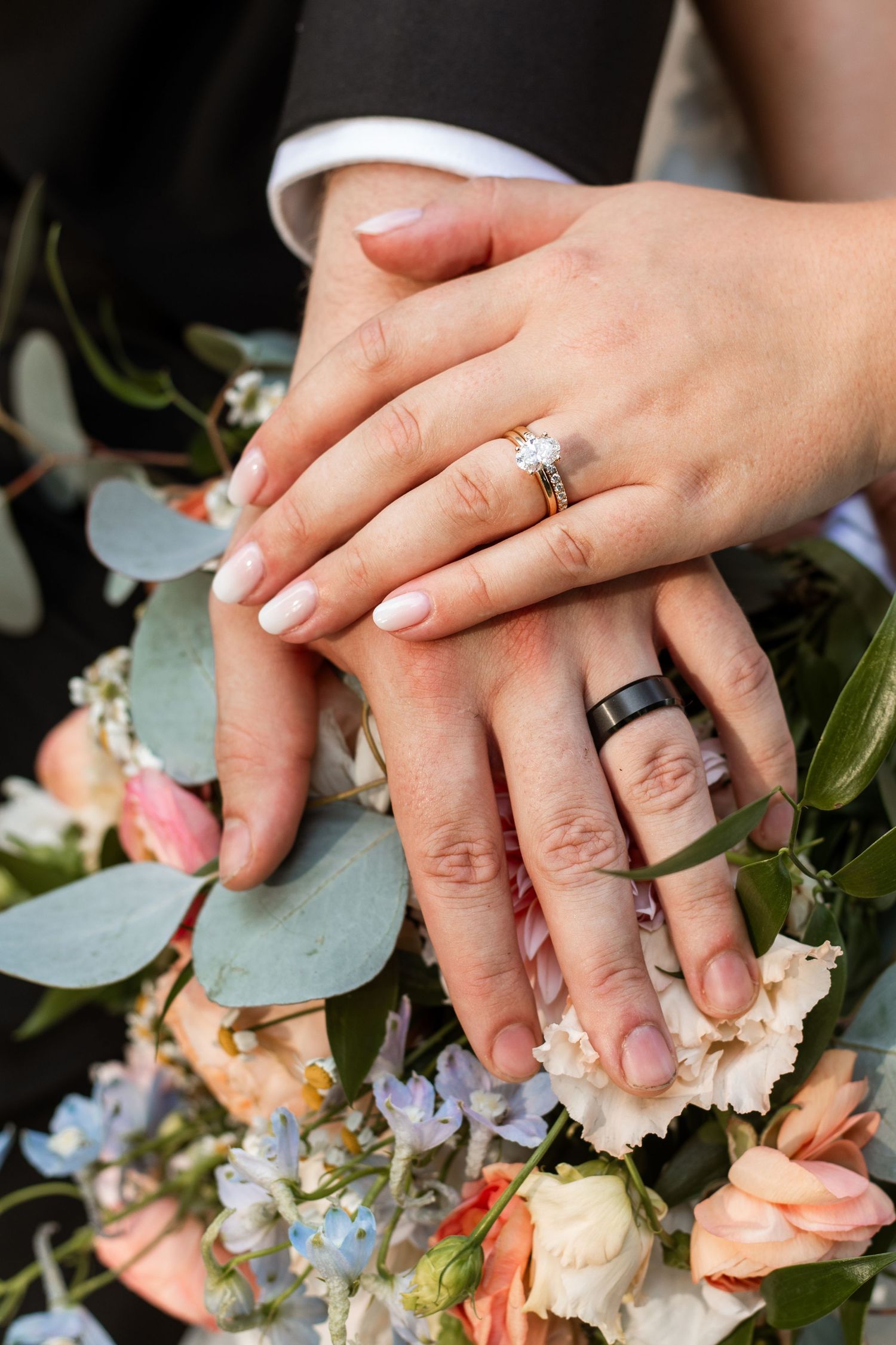 Two hands with wedding rings rest on a romantic bouquet of peach roses, blue flowers and eucalyptus leaves.