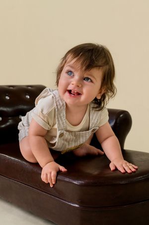 Happy toddler in striped overalls crawls on brown leather couch while smiling brightly.