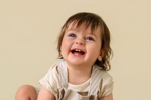 A laughing baby with dark hair wearing a white outfit against a neutral beige background.