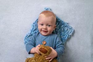 Baby wearing blue sweater plays with soft brown teddy bear while sitting on blue blanket.