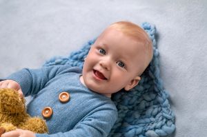 Baby in light blue outfit lies on soft blue blanket with plush brown teddy bear.