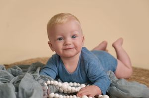 A baby in a blue onesie lies on beige carpet while playing with white cotton balls.