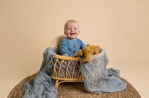 Baby wearing blue outfit sits in woven basket with gray blanket and brown teddy bear.