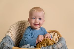 A happy baby wearing a blue sweater sits in a wicker chair with a blanket.