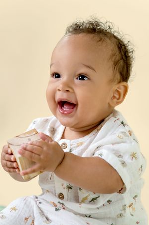 Happy baby wearing white floral outfit laughs joyfully while sitting against beige background.